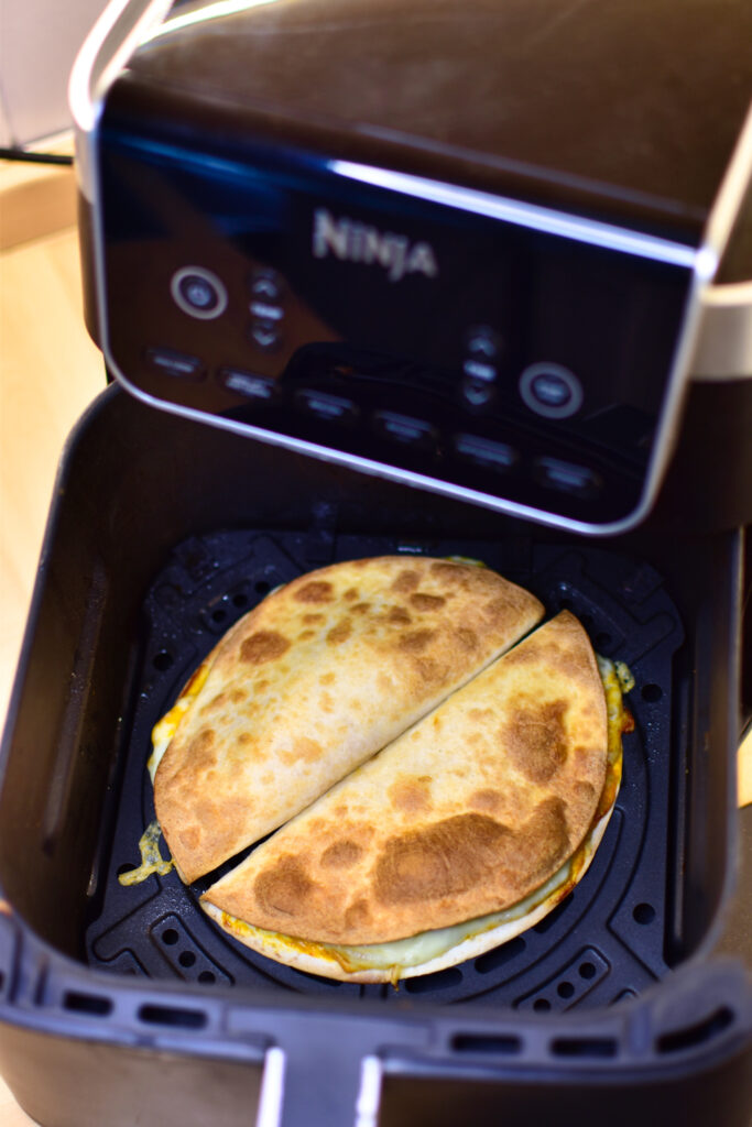 Zwei gefüllte und geklappte Tortilla-Fladen liegen nebeneinander im Korb eines Airfryers kurz vor dem Backvorgang.