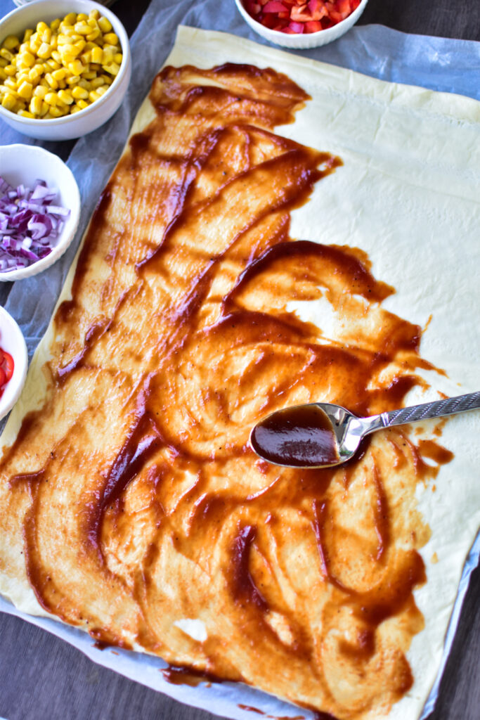 Der fertige Pizzateig wird mit Sauce bestrichen. Daneben stehen die einzelnen Zutaten in Schälchen.