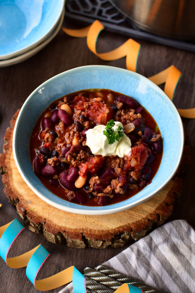 Partytisch mit einer blauen Schale Chili con Carne, serviert mit Toppings wie Sauerrahm und Petersilie.