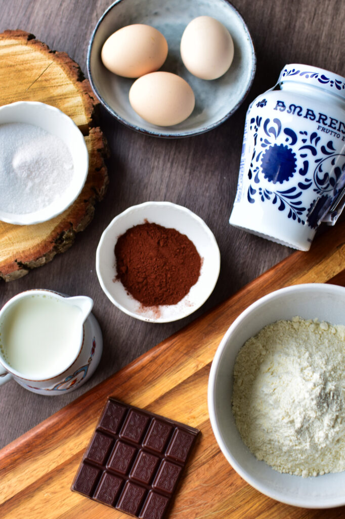 Alle zutaten auf einen Blick: Eier, Zucker, Kakao, Milch, Mehl, Schokolade und Fabbri Amarenakirschen auf einem Holztisch.