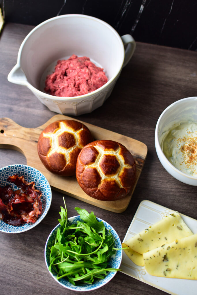 Alle Zutaten auf einen Blick. In der Mitte knusprige Laugenbrötchen auf einem Holzbrett.