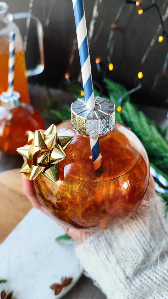 Nahaufnahme eines funkelnden goldenen Punschs, mit glitzerndem Effekt und schwimmenden Anissternen, serviert in einem Glas in Baumkugelform – festliche Tischdekoration zu Weihnachten.