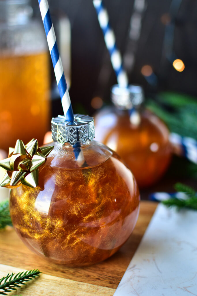 Heißer alkoholfreier Goldpunsch mit Apfelsaft, weißem Tee und essbarem Goldglitzer – elegant serviert in runden Weihnachtskugelgläsern mit festlicher Beleuchtung im Hintergrund.