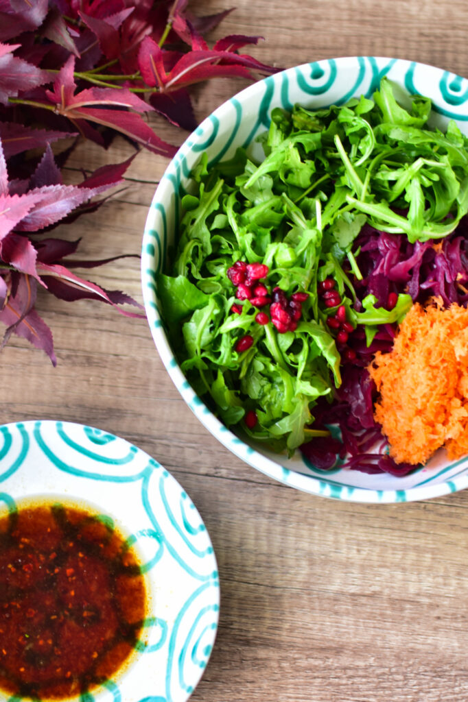 Bunter Salat mit Rucola, Möhren, Rotkraut und Granatapfelkernen in einer großen grünen Schüssel. Das Dressig mit Sojasauce steht daneben.
