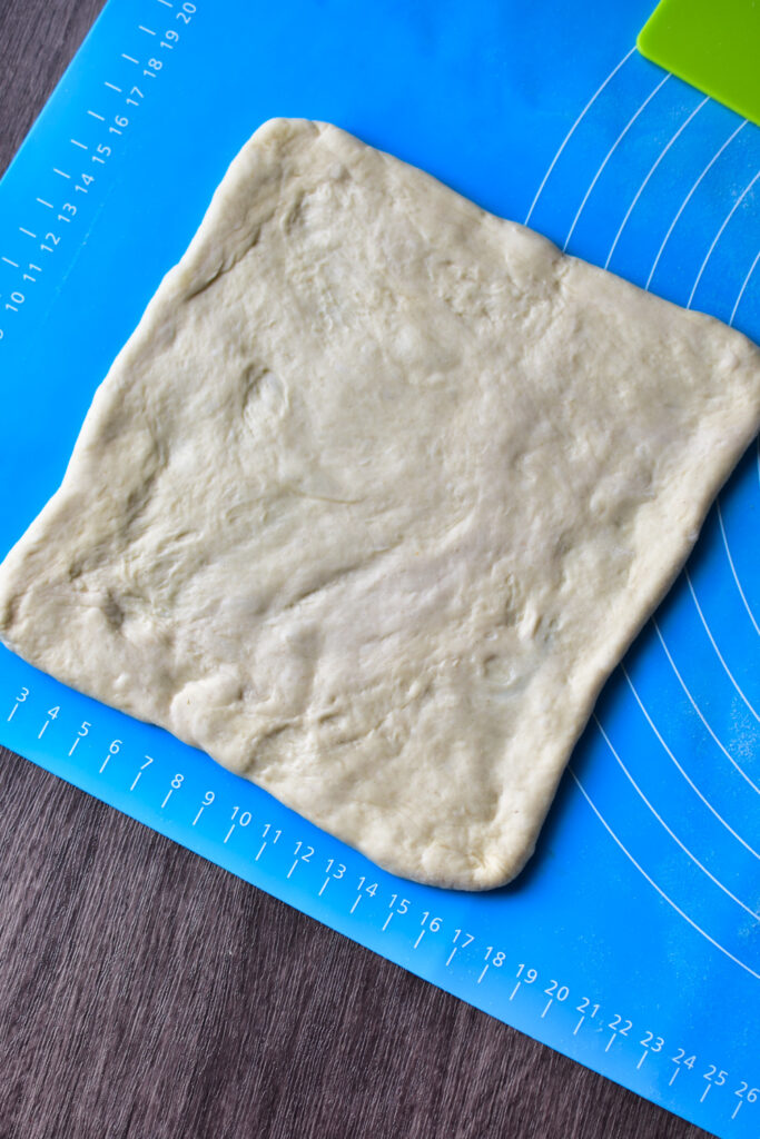 Teig wird auf einer Backmatte zu einem Quadrat ausgerollt.