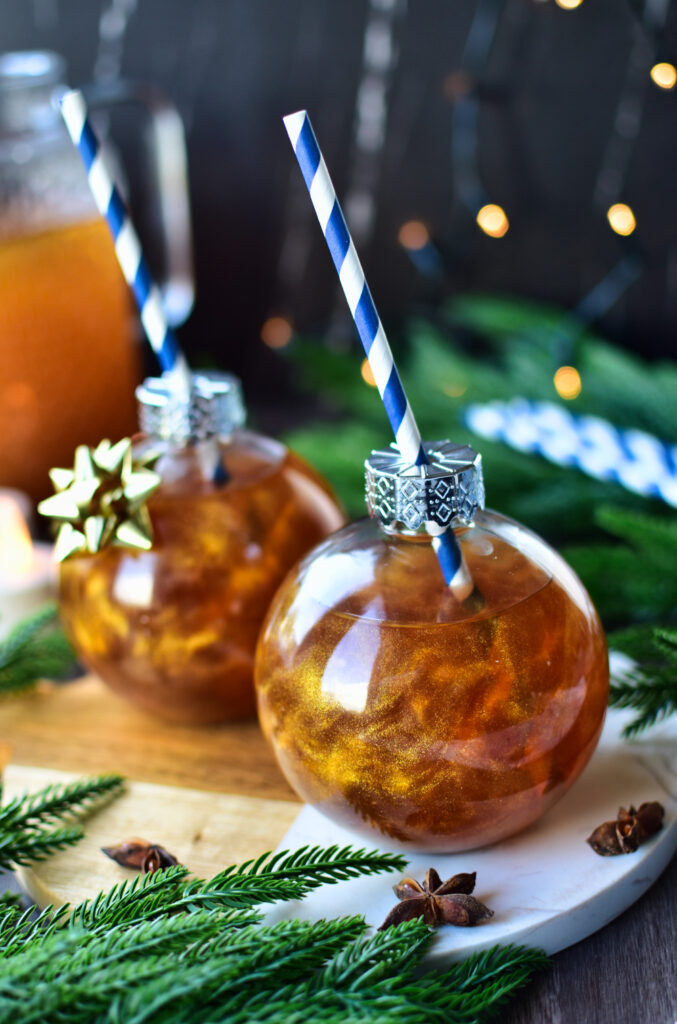Weihnachtlicher goldener Punsch in glitzernden Baumkugel-Gläsern mit Trinkhalm, serviert auf einem Holztisch mit Zimtstangen, Anissternen und Tannenzweigen als Dekoration.