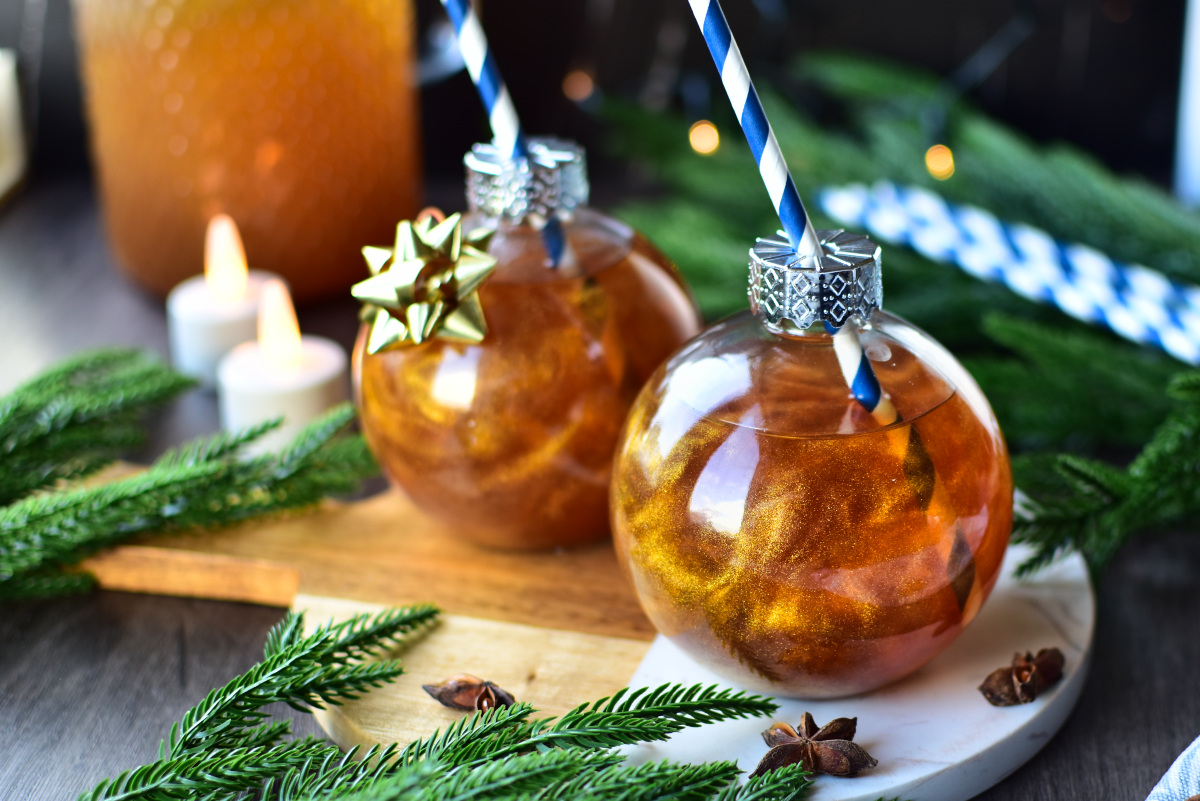 Selbstgemachter goldener Weihnachtspunsch mit Apfel, Zimt und Anis, liebevoll angerichtet in Seaside No.64 Baumkugel-Gläsern mit Strohhalm und goldenen Akzenten.