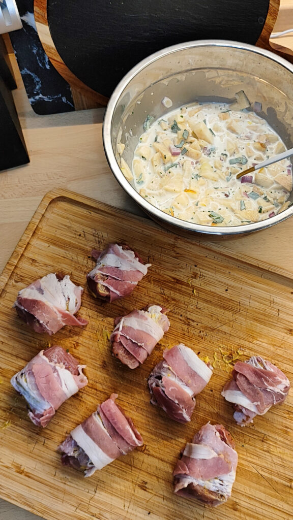 Vorbereitung. Die Filets liegen auf einem Holzbrett und sind mit Bacon umwickelt. Daneben die Apfel-Sahne-Sauce in einer Schüssel.