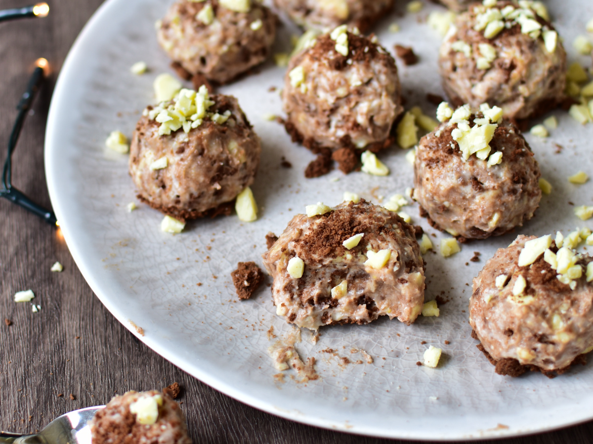 Cremige Cheesecake-Bällchen mit Spekulatius und geraspelter weißer Schokolade – weihnachtliches Dessert ohne Backen auf einem Holzbrett angerichtet.