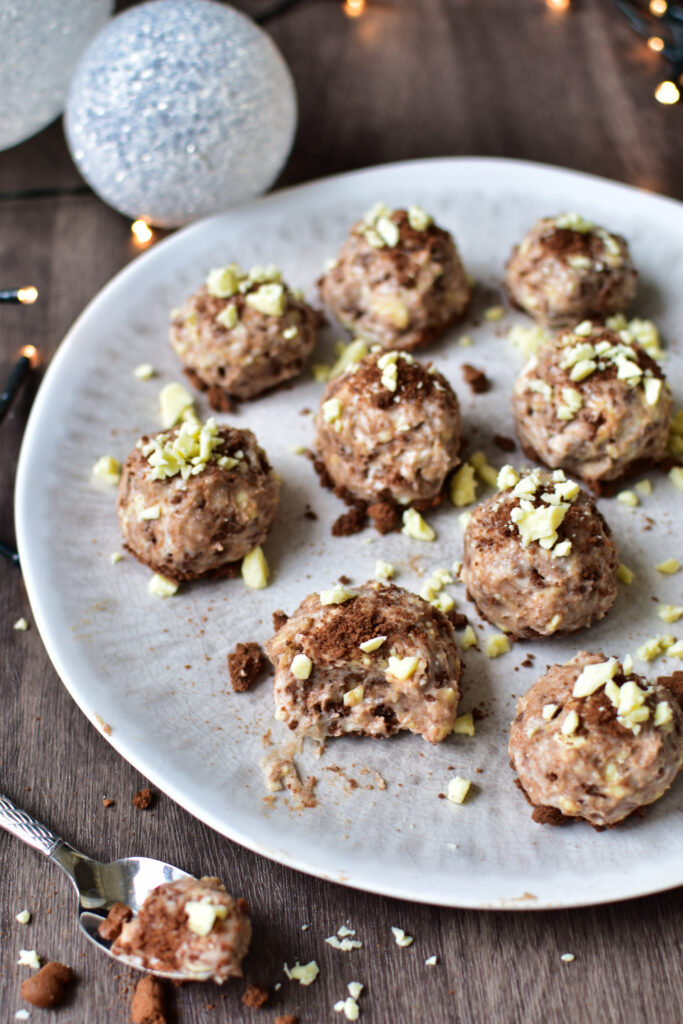 Käsekuchen-Bällchen mit Spekulatiusbröseln und Kakaopulver bestreut, dekorativ auf rustikalem Teller mit Weihnachtslichtern.