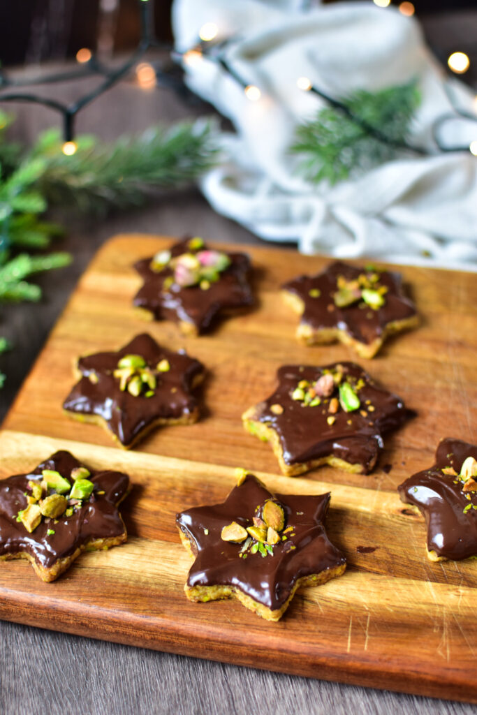 Pistazien Sterne mit Schokoglasur auf einem Holzbrett Tannenzweige und Weihnachtslichter im Hintergrund.