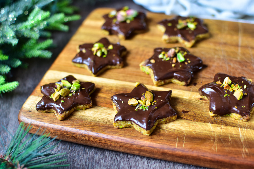 Schoko Pistazien Plätzchen auf einem Brett mit weihnachtlichen Tannenzweigen im Hintergrund.