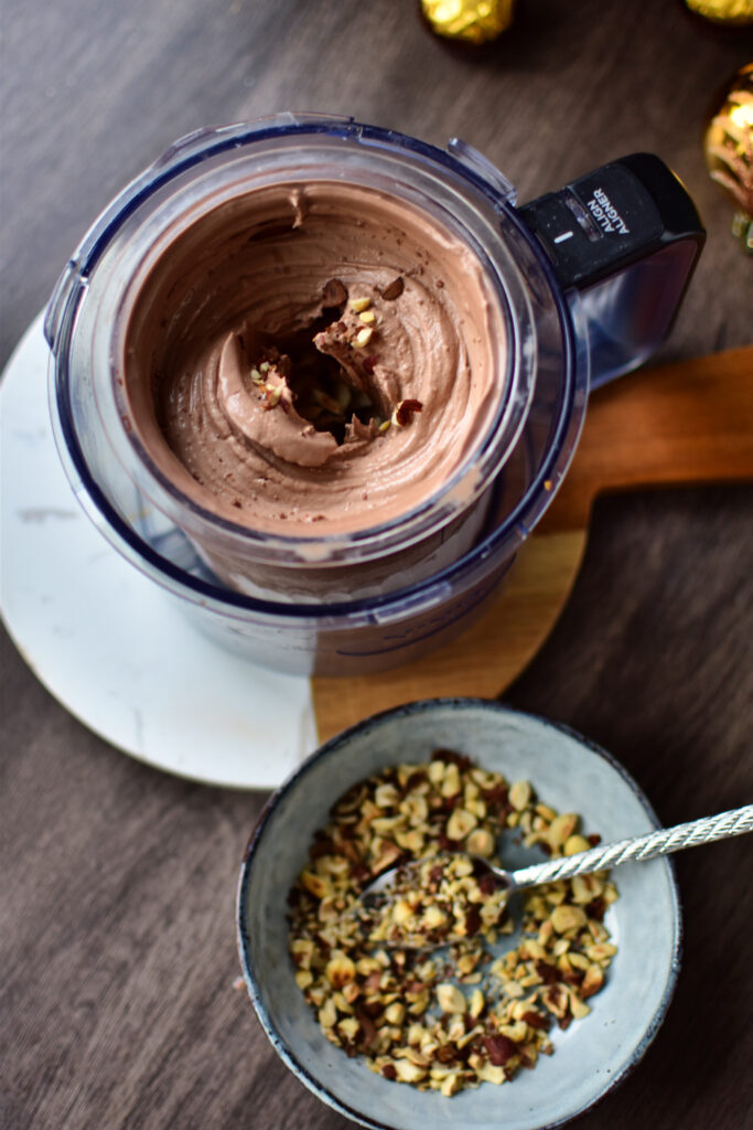 Cremiges Schokoeis mit gerösteten Haselnüssen vor dem Mix-In Programm des Ninja Creami mit einem Loch in der Mitte, in das die Haselnuss-Stückchen eingefüllt werden.