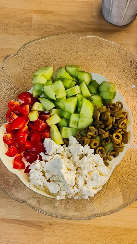Frische Tomaten, Gurken und Oliven vermischt mit einem cremigen Joghurt-Senf-Dressing in einer großen weißen Salatschüssel.
