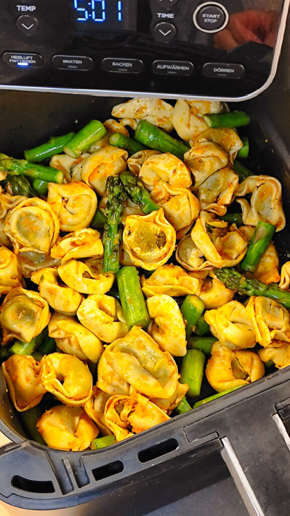 Goldbraun gebackene, knusprige Tortellini und geröstete grüne Spargelstücke direkt nach dem Backen im Korb des Airfryers.
