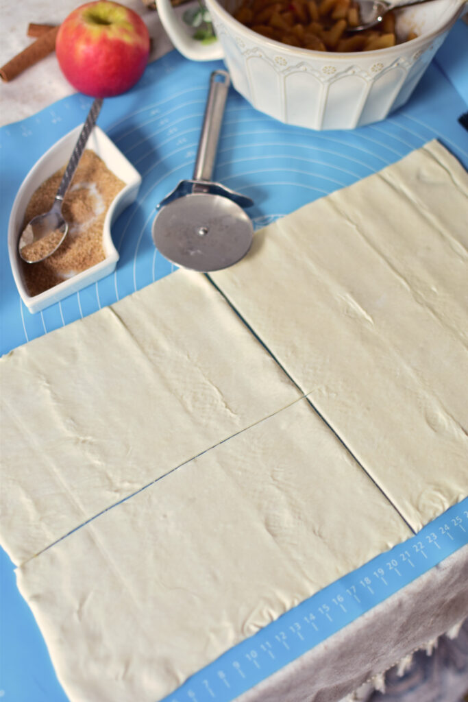 Blätterteig wird in Rechtecke geschnitten. Er liegt ausgebreitet auf einer blauen Backmatte, daneben die Zutaten für die Apfeltaschen und ein Pizzaschneider.
