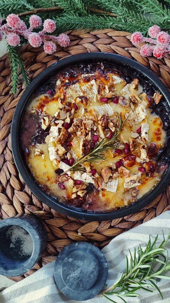 Zerlaufener Camembert aus der Heißluftfritteuse mit gerösteten Walnüssen und frischem Rosmarin in einer schwarzen Auflaufform. Drumherum befindet sich weihnachtliche Deko mit Tannenzweigen und roten Cranberries.