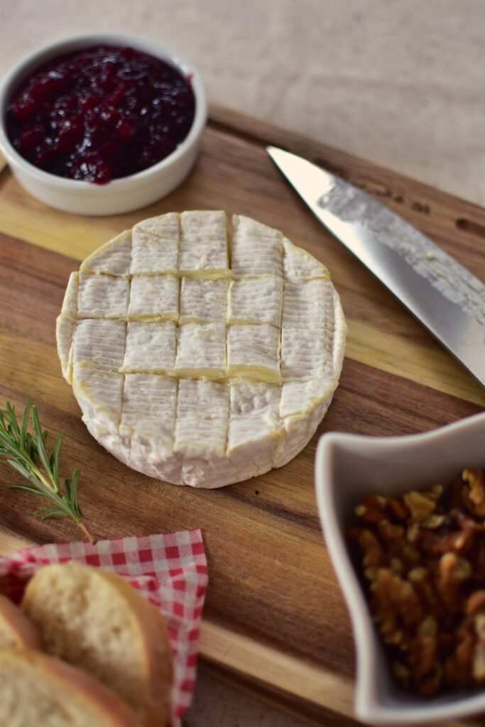 Camembert auf einem Schneidebrett mit einem eingeschnittenen Muster auf der Oberseite. Daneben ein Messer und eine Schale mit Preiselbeersauce.